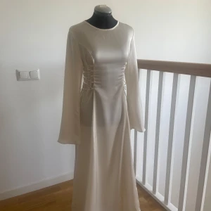 Ljus långklänning med snörning i satin - Elegant ljus långklänning i satin med vida långa ärmar och snygg snörning i midjan på sidorna. Klänningen har en rund halsringning och ett stilrent, minimalistiskt uttryck. Perfekt för dig som vill sticka ut med en drömmig och modern look.