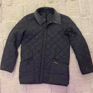Quiltad Barbour jacka med manchesterkrage - Mörkblå quiltad jacka från Barbour med klassisk manchesterkrage i grått. Jackan har tryckknappar framtill, två stora fickor och Barbour-logga på ena fickan. Perfekt för dig som gillar stilren och tidlös design.