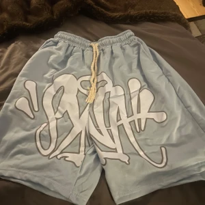 Syna world Ljusblå shorts med graffiti-tryck  - Ljusblå syna world shorts med stort vitt graffiti-inspirerat tryck framtill. Materialet är mjukt och lätt, perfekt för varma dagar. Tröjan kan köpas som set med shortsen eller separat. Priset kan diskuteras vid snabb affär.