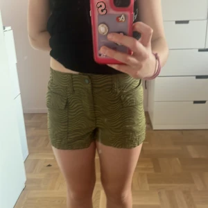 Gröna mönstrade shorts med fickor - Snygga gröna shorts med vågigt zebramönster och praktiska fickor framtill. De har midwaist-passform, bälteshällor och knappstängning. Perfekta för varma dagar och ger en cool vibe till din outfit.