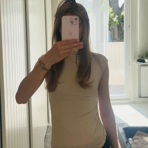 Beige polotopp från Gina Tricot - Säljer en beige polotopp från Gina Tricot med ärmlös design och tight passform. Toppen har hög hals och snygg rynkdetalj på båda sidor. 