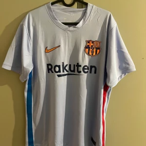 FC Barcelona bortatröja Nike - Snygg ljusblå FC Barcelona bortatröja från Nike med klubbmärke och Rakuten-logga på bröstet. Tröjan har korta ärmar och detaljer i blått och rött på sidorna. Perfekt för dig som älskar Barca och vill visa ditt stöd på matchdagar.