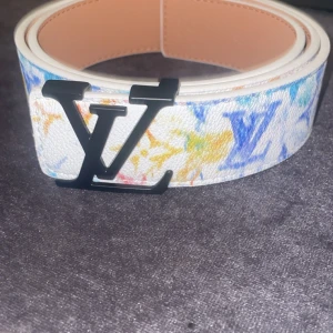 Louis Vuitton färgglatt skärp med LV-spänne - Louis Vuitton bälte i vitt skinn med färgglada inslag i blått, gult och orange. Bältet har ett stort svart LV-spänne och klassiskt monogrammönster i olika färger. Perfekt statement piece för att lyfta din outfit.