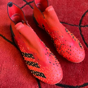 Adidas Predator röda fotbollsskor - Säljer ett par röda Adidas Predator fotbollsskor med svarta detaljer och ikoniska tre ränder på sidan. Skorna har en strumpliknande passform utan snörning och gummitaggar på ovandelen för extra grepp på bollen. Storlek 41 1/2 Saknar snören  Nypris: 2000kr Pris kan diskuteras.