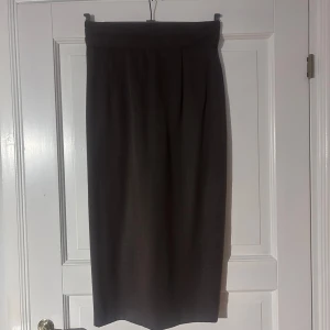 Mörkbrun långkjol i polyester - Snygg mörkbrun långkjol med hög midja och diskreta veck framtill. Kjolen har en klassisk rak passform och stängs med knapp och dragkedja bak. Tillverkad i en mjuk blandning av polyester och viskos, perfekt för en stilren look.