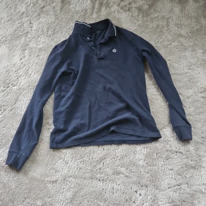 Mörkblå långärmad pikétröja - Snygg mörkblå långärmad pikétröja från WESC med klassisk krage och tre knappar framtill. Tröjan har en vit broderad logga på bröstet och vit detalj på kragen. Perfekt för en stilren och avslappnad look.