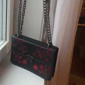 Svart axelväska med röda broderade blommor - Svart axelväska i skinnimitation med grov silverkedja som axelrem. Väskan har broderade röda blommor på framsidan och ett snyggt metallspänne. 