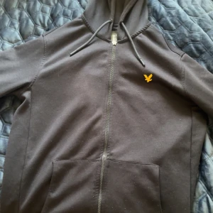  hoodie från Lyle & Scott - Svart/marin blå hoodie från Lyle & Scott med dragkedja och klassisk gul logga på bröstet. Tröjan har huva med snörning och känguruficka framtill. Perfekt för en chill och stilren look.