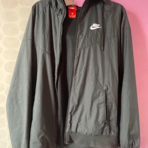 Svart Nike vindjacka med huva - Svart vindjacka från Nike med vit logga på bröstet. Jackan har huva, dragkedja framtill och två fickor med dragkedja. Lätt och sportig känsla med meshfoder på insidan. Perfekt för dig som gillar enkel och stilren streetwear.