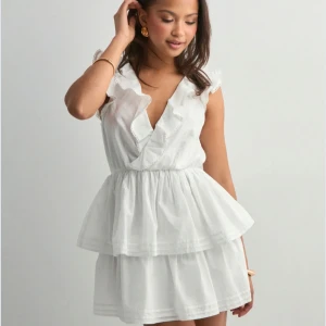 Vit volangklänning från Nelly XXS perfect frill dress - Supersöt vit kortklänning från Nelly med volanger, djup v-ringning och öppen rygg med knyt. Klänningen har dubbla lager i kjolen och är ärmlös med spetsdetaljer längs volangerna. Perfekt för sommarens alla tillfällen! Helt ny med prislapp