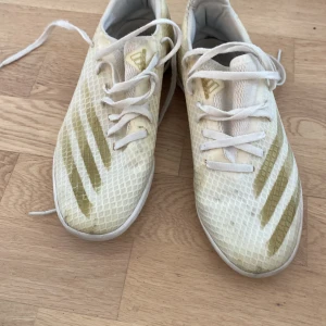 Vita Adidas fotbollsskor strl 37⅓ - Säljer ett par vita Adidas fotbollsskor med guldiga detaljer och snörning. Skorna har ett mönstrat mesh-överdrag och platt sula som ger bra grepp på planen. Perfekta för dig som vill sticka ut med stilrena och sportiga skor.