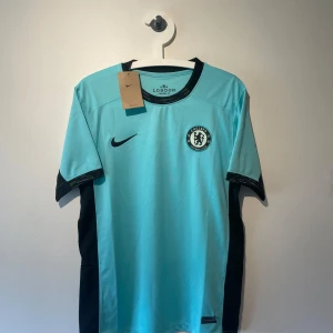 Chelsea FC Nike fotbollströja  - Chelsea FC fotbollströja från Nike i turkosblå färg med svarta detaljer på sidor och krage. Broderat Chelsea-märke på bröstet och Nike-logga. Tillverkad i lätt och ventilerande Dri-FIT-material, perfekt för träning eller match.