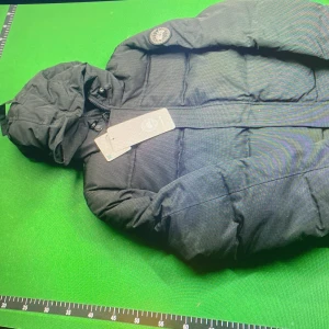 Svart pufferjacka med huva - Snygg svart pufferjacka från canada goose med huva och dragkedja framtill. Jackan har quiltad design och en patch på ena ärmen. Perfekt för kalla dagar när du vill hålla stilen och värmen.