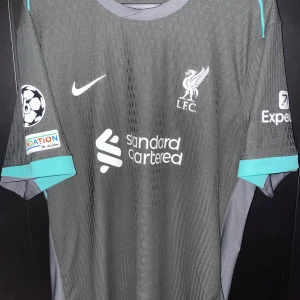 Liverpool Champions League Alexander-Arnold Nike Authentic/Vaportröja XXL - Liverpool FC Champions League matchtröja från Nike med Alexander-Arnold och nummer 66 på ryggen. Tröjan är mörkgrå med turkosa detaljer vid ärmslut och axlar, samt vita tryck och loggor. Tillverkad i Dri-FIT material som andas, perfekt för fotboll eller träning.