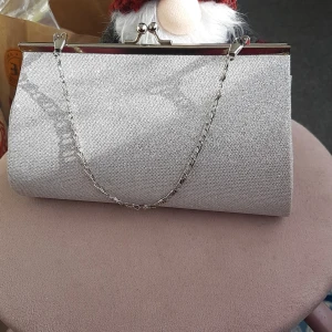 Glittrig silver kuvertväska med kedja - Snygg kuvertväska i glittrigt silver med metallkedja och klassiskt klolås. Perfekt att bära i handen eller över axeln. Stilren och enkel design som ändå sticker ut med sitt glittrande tyg och metalliska detaljer.