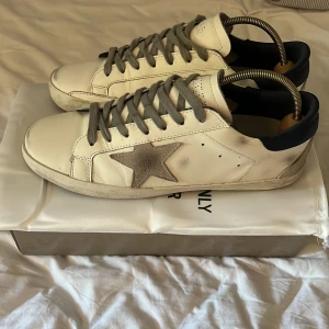 Golden Goose Superstar sneakers vit/grå - Golden Goose Superstar sneakers i vitt skinn med grå mockastjärna på sidan, grå snörning och mörkblå häl. Klassisk rund tå och platt sula. Snyggt slitna detaljer för en cool streetstil. Kommer med originalpåse och låda.