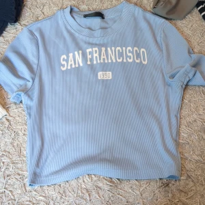 Baby tee - Snygg ljusblå ribbad t-shirt med trycket 'San Francisco 1990' i vitt framtill. T-shirten har korta ärmar och en relaxed fit, perfekt för en chill och trendig look. Mjuk och skön bomullskänsla.