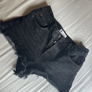 Svarta jeansshorts med fransig kant - Svarta jeansshorts från Zara med hög midja och rå, fransig kant nertill. Klassisk femficksmodell med knappgylf och slitna detaljer för en cool, avslappnad vibe. Perfekta till sommaren och lätta att matcha med allt.