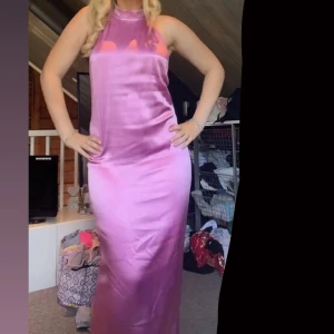 Lång rosa satinklänning med öppen rygg - Säljer en lång, glansig rosa klänning i satin med halterneck och smala axelband. Klänningen har öppen rygg, slits bak och en figurnära passform. Perfekt för dig som vill sticka ut med en elegant och trendig look. Använd 1 gång!