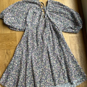 Blommig kortklänning med puffärm - Supersöt kortklänning med blommigt mönster i lila, vit, gul och grön. Klänningen har v-ringning med snörning i ryggen och stora puffärmar. Den gör sig inte riktigt rättvis på bild, kan skicka bild på om det är utav intresse.