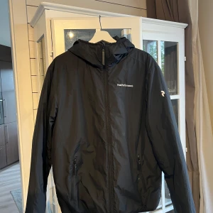 Vadderad jacka Peak Performance  - Svart jacka från Peak Performance i storlek large herrstorlek. Jackan har huva, hel dragkedja framtill och två fickor med dragkedja. Snygg minimalistisk design med logga på bröstet och ärmen. Perfekt för blåsiga dagar och enkel att matcha med allt. Modellen funkar för både dam och herr. 