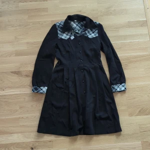 Vintage Svart skjortklänning med rutiga detaljer - Vintage svart skjortklänning med knappar framtill och rutiga detaljer i vitt och grått på axlar och ärmslut. Klänningen har lång ärm, klassisk krage och lätt utställd kjol.