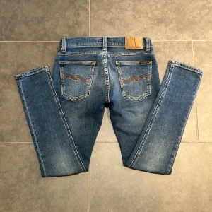 Blå jeans från Nudie Jeans Co - Snygga blå jeans från Nudie Jeans Co med klassisk femficksdesign och kontrastsömmar. Jeansen har en rak passform och coola broderade detaljer på bakfickorna. Perfekta för dig som gillar en tidlös och avslappnad stil. Modellen heter tight terry och sitter väldigt bra. Storlek 28/30. Ny pris: 1600kr. Priset går lätt att ta ner så fråga vad ni vill!