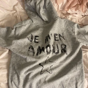 Grå hoodie med tryck och stjärna - Säljer en grå hoodie med svart texttryck 'Je m'en amour' och en stjärna på bröstet. Tröjan har huva med snörning och är långärmad. Perfekt för en chill och avslappnad stil.