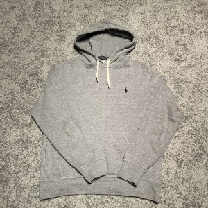 Grå hoodie från Ralph Lauren - Säljer en stilren grå hoodie från Ralph Lauren med det klassiska broderade logotypen på bröstet. Tröjan har en justerbar huva med vita snören och en praktisk magficka. Perfekt för en avslappnad look.