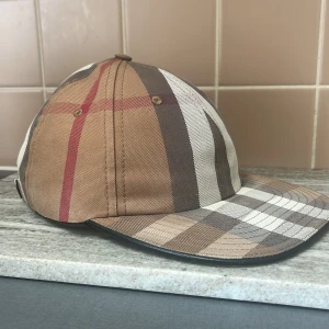 Rutig keps från Burberry - Snygg rutig keps från Burberry i jordnära toner med klassiskt mönster. Kepsen har justerbart spänne baktill och broderad logotyp. Perfekt för en stilren look.