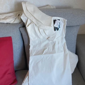 Vit klänning med volangdetaljer - Elegant vit klänning med volangdetaljer och en asymmetrisk design. Perfekt för sommarens festligheter. Klänningen har en oneshoulder-stil som ger en modern touch. Mycket stretch passar även M.