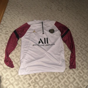 Vit och vinröd fotbollströja från Paris Saint-Germain - Säljer en snygg vit och vinröd långärmad fotbollströja från Paris Saint-Germain med Jordan-logga. Tröjan har klubbens emblem på bröstet och texten 'Accor Live Limitless' på framsidan. Perfekt för fans av PSG!