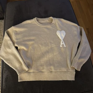 Beige stickad tröja med hjärtdetalj - Säljer en stilren beige stickad tröja med en vit hjärtdetalj och bokstaven 'A' på bröstet. Tröjan har långa ärmar och ribbade muddar vid ärmslut och nederkant. Perfekt för en mysig dag!
