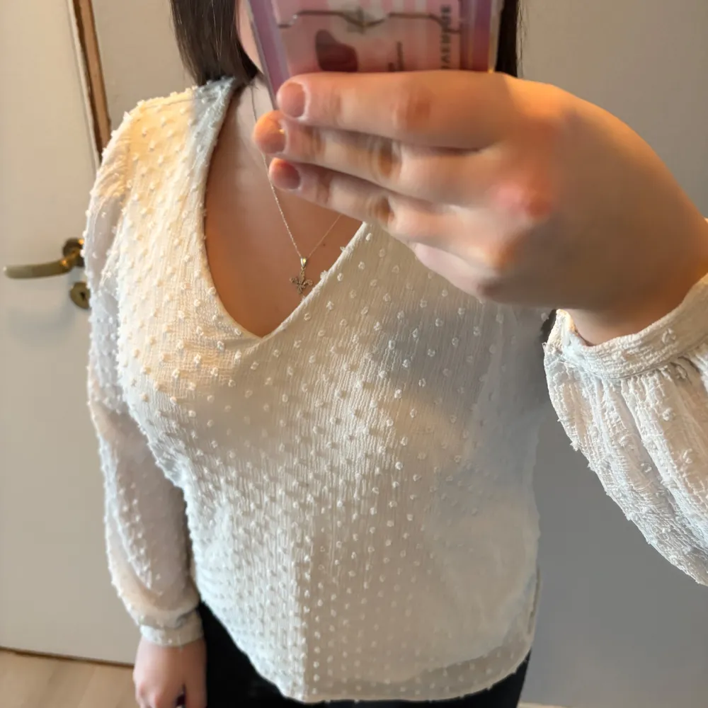 Säljer en elegant vit blus med v-ringning och långa ärmar. Blusen har ett diskret prickigt mönster och knappar vid ärmsluten. Perfekt för en stilren look.. Puserot.
