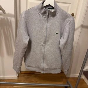 Grå zip-tröja från Lacoste - Säljer en stilren grå zip-tröja från Lacoste med dragkedja och hög krage. Tröjan har långa ärmar och det klassiska Lacoste-logotypen på bröstet. Perfekt för en avslappnad stil. Storlek S/M. Nypris: 1600