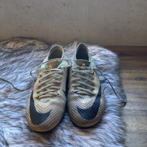 Nike Mercurial fotbollsskor - Säljer ett par Nike Mercurial fotbollsskor i beige och svart. Skorna har en snygg design med snörning och är perfekta för fotbollsplanen. De har en spetsig tå och är tillverkade i syntetmaterial för bästa prestanda.(konsgräsdobbar)