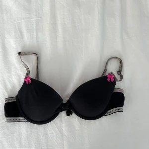 Svart och rosa bh från Juicy Couture - Snygg svart bh från Juicy Couture med rosa insida och dekorativa rosa rosetter. Justerbara axelband.