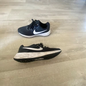 Svarta Nike sneakers med vit sula - Snygga svarta Nike sneakers med vit sula och klassisk vit swoosh-logga på sidan. Skorna har snörning och en bekväm passform, perfekta för både träning och vardag. Den svarta ovandelen är i syntetmaterial med en stilren design.