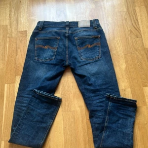 Mörkblå Nudie Jeans - Säljer ett par snygga mörkblå jeans från Nudie Jeans. De har en klassisk femficksdesign med orange sömmar och en bekväm passform. Perfekta för en stilren look.