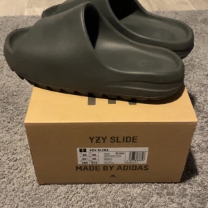 Mörk gråa YZY Slide från Adidas - Säljer ett par mörk gråa YZY Slide från Adidas. Dessa slides har en stilren design med en bred rem och räfflad sula för extra grepp. Perfekta för en avslappnad stil.