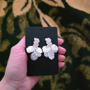Vita örhängen med kristaller - Eleganta vita örhängen med blommiga detaljer och glittrande kristaller. Perfekta för att ge en lyxig touch till din outfit. De har en unik design med pärlemorskimrande blommor och rektangulära kristaller som ger en vacker glans.