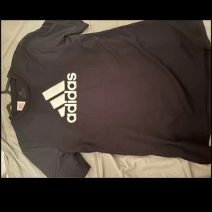 Svart t-shirt från Adidas - Säljer en svart t-shirt från Adidas med deras ikoniska logga i vitt på bröstet. T-shirten har en klassisk design med korta ärmar och rund hals. Perfekt för en sportig look eller avslappnad stil.