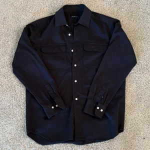 Overshirt från Riley - Snygg svart långärmad skjorta från Riley med klassisk krage och vita knappar. Skjortan har två bröstfickor med lock och en stilren design som passar perfekt för olika tillfällen.