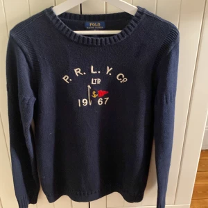 Mörkblå tröja från Ralph Lauren - Säljer en snygg mörkblå tröja från Ralph Lauren med broderad text och ett litet rött flaggmotiv på framsidan. Tröjan har långa ärmar och en rund halsringning. Perfekt för en stilren look. Tröjan är storlek XL barn vilket passar som Small.