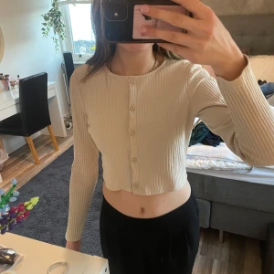 Beige ribbad kofta - Säljer en stilren beige ribbad kofta med knappar framtill. Den är croppad och har långa ärmar, perfekt för en trendig look. Passar bra till både jeans och kjol.