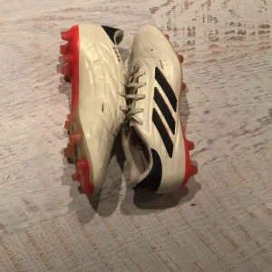 Vita fotbollsskor från Adidas - Säljer ett par vita Adidas fotbollsskor med svarta detaljer och röda dobbar. Skorna har snörning och är perfekta för fotbollsplanen. De är tillverkade i syntetmaterial och har en klassisk design med tre svarta ränder på sidan.