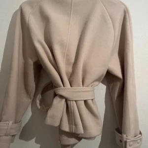 Beige kappa från Nelly - Snygg beige kappa från Nelly med knytbälte i midjan och klassisk krage. Jackan har långa ärmar med justerbara spännen vid ärmsluten. Perfekt för en stilren look, använt bara 3 gånger helt ny 