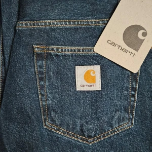 Blå jeansbyxor från Carhartt landon  - Säljer ett par klassiska blå jeansbyxor från Carhartt. Byxorna har en regular waist och en loose fit, perfekt för en avslappnad stil. De är tillverkade i slitstarkt denim och har en dragkedja framtill. Carhartt-loggan är snyggt placerad på bakfickan.