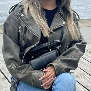 Olivgrön skinnjacka med dragkedja - Snygg olivgrön skinnjacka med dragkedja och axelremmar. Jackan har en cool bikerstil med detaljer som knappar och en asymmetrisk dragkedja framtill. Perfekt för en trendig look. storleken är M, men passar även i S