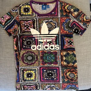 Färgglad t-shirt från Adidas - Säljer en färgglad t-shirt från Adidas med ett unikt mönster i olika färger och stor logga på framsidan. T-shirten har korta ärmar och en rund halsringning. Perfekt för dig som vill sticka ut med en cool och livfull stil!